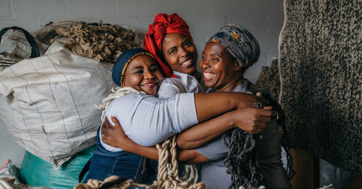 Female weavers who create Coral & Hive rugs together in front of wool and weaving looms. Coral & Hive rugs are available at Sarza home goods, furniture and rugs store.